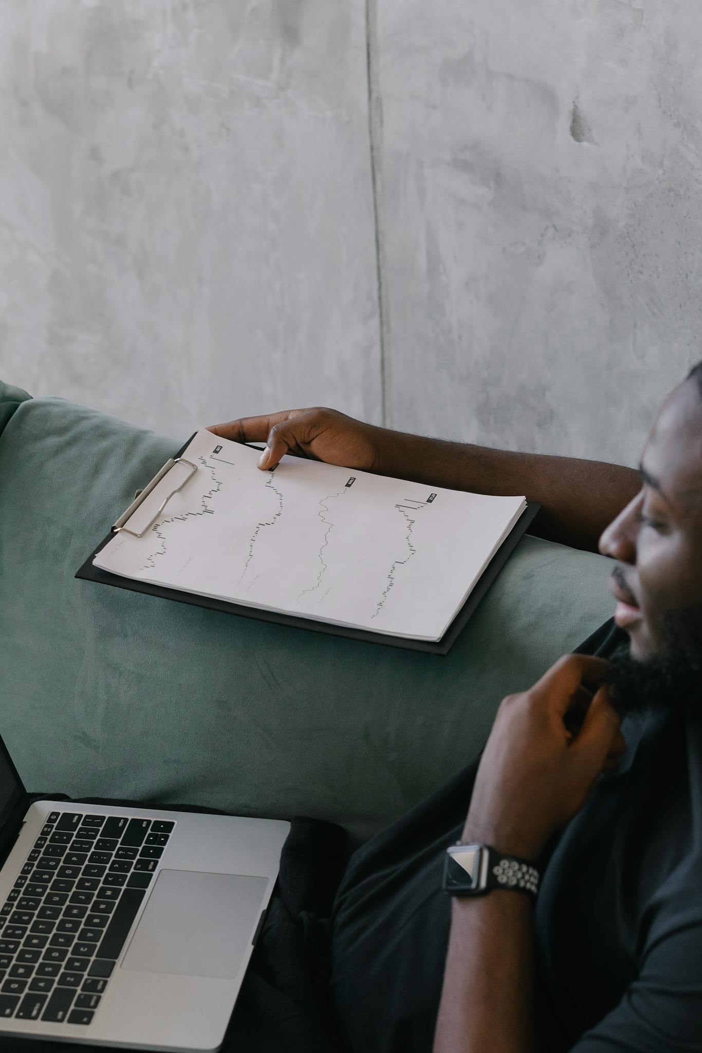 Focused businessman in black shirt reviews financial graphs on clipboard beside laptop. Concrete wall background.